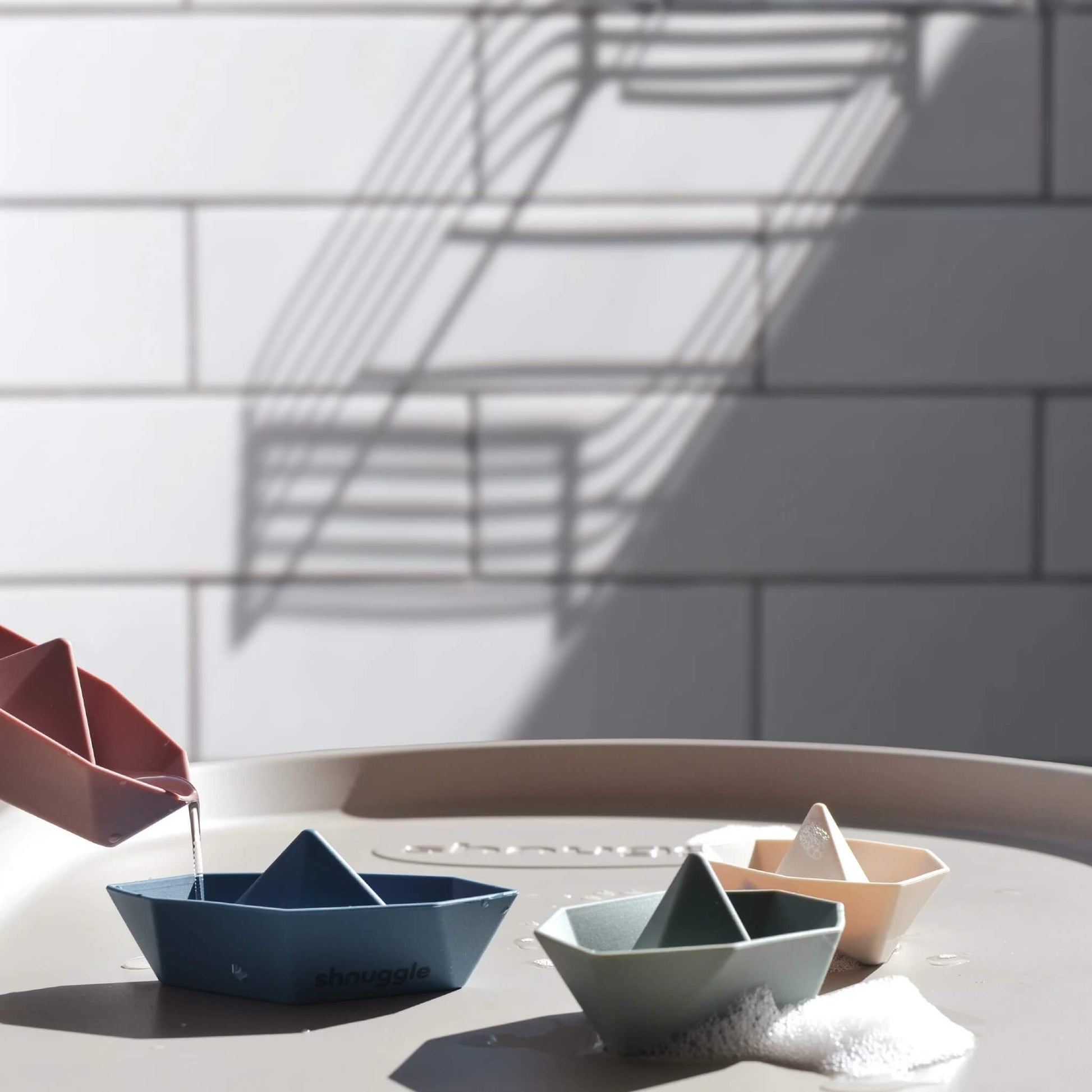  Water being poured from one toy boat into another, with several geometric boat toys resting on a baby changing mat  with a tiled wall in the background.