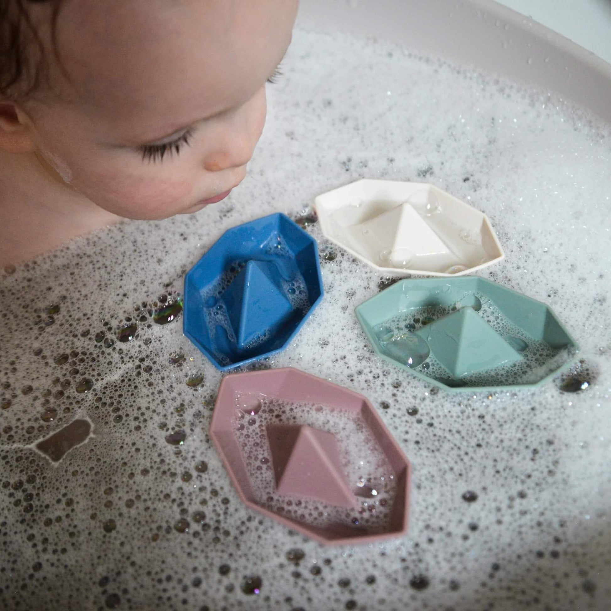A close-up of a baby leaning over bath water, watching several small boat toys floating among bubbles.
