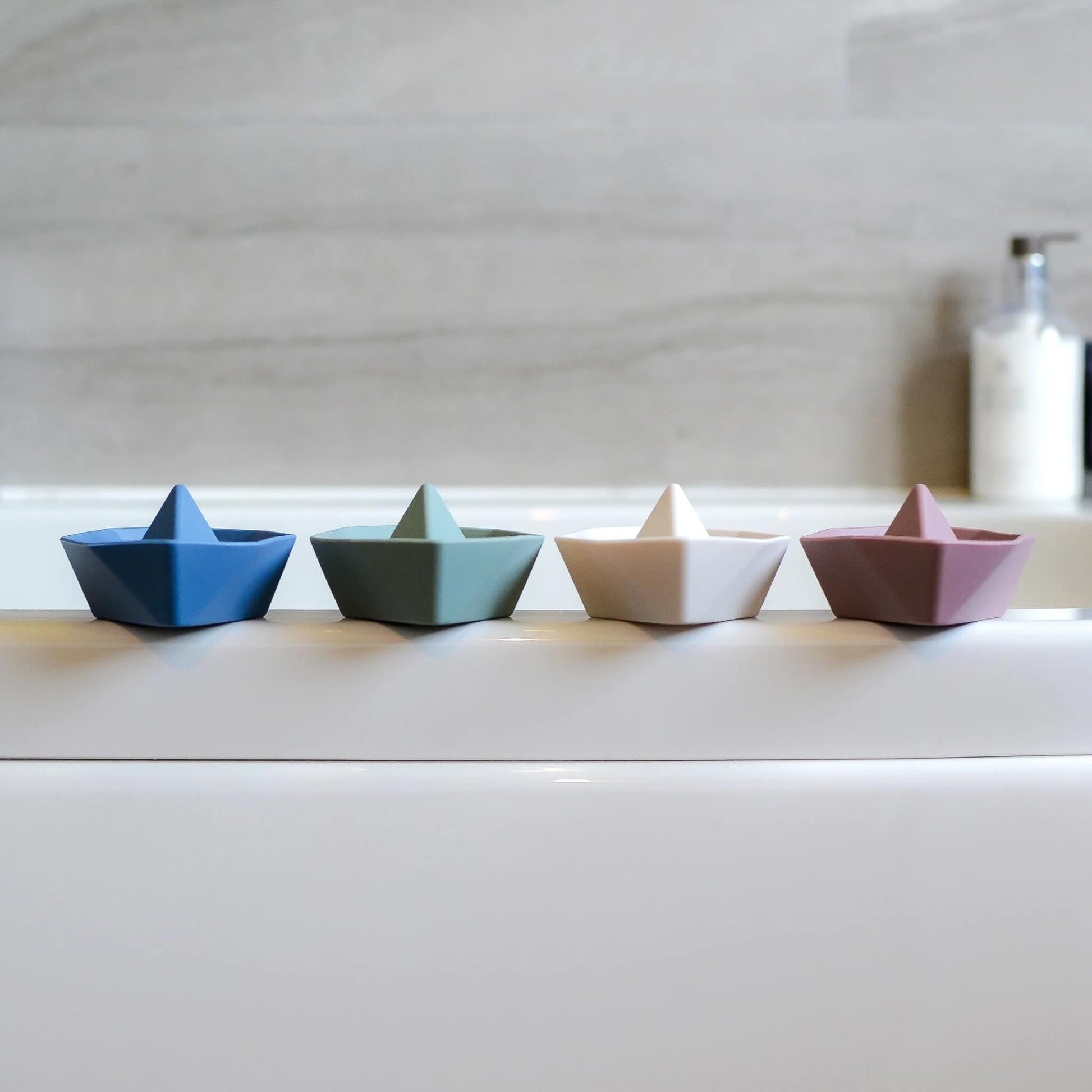 A row of small boat toys lined up along the edge of a bath, shown in different muted colours.