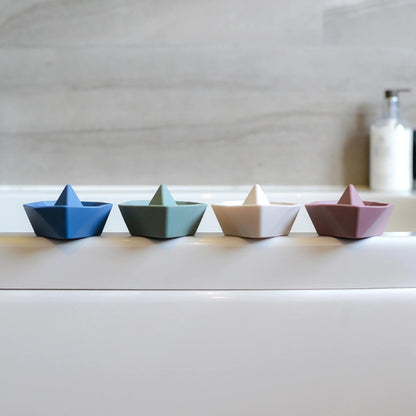 A row of small boat toys lined up along the edge of a bath, shown in different muted colours.