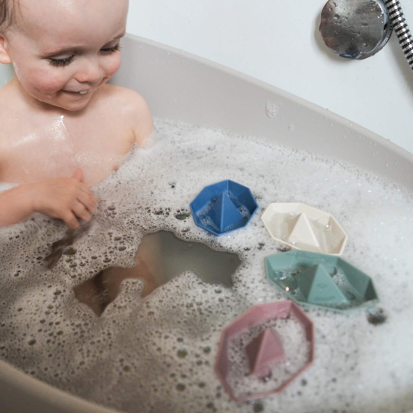 A baby sitting in a bath filled with bubbles, watching bath boats float on the water during play.
