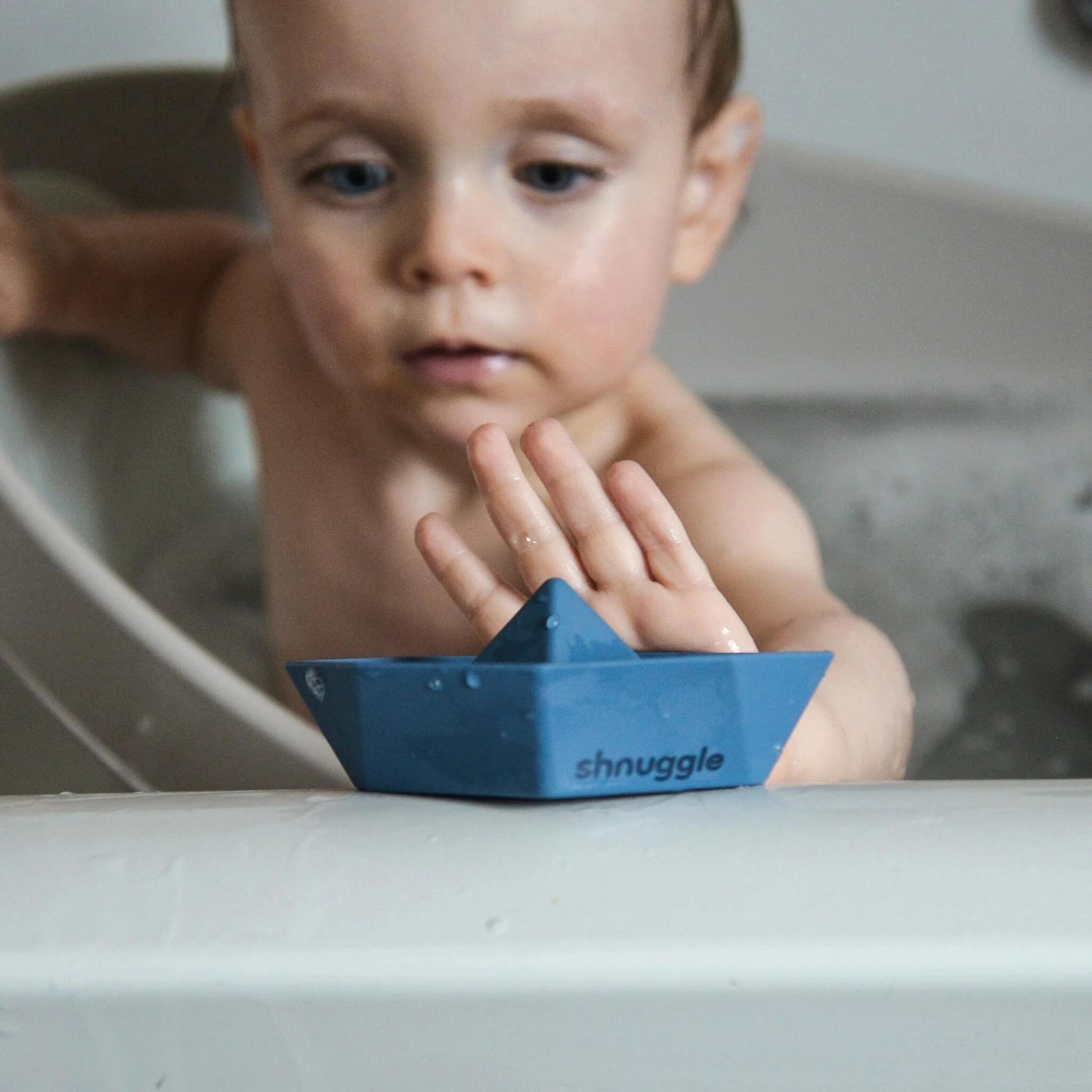 A baby reaching towards a blue bath boat resting on a bath edge, showing scale during use.