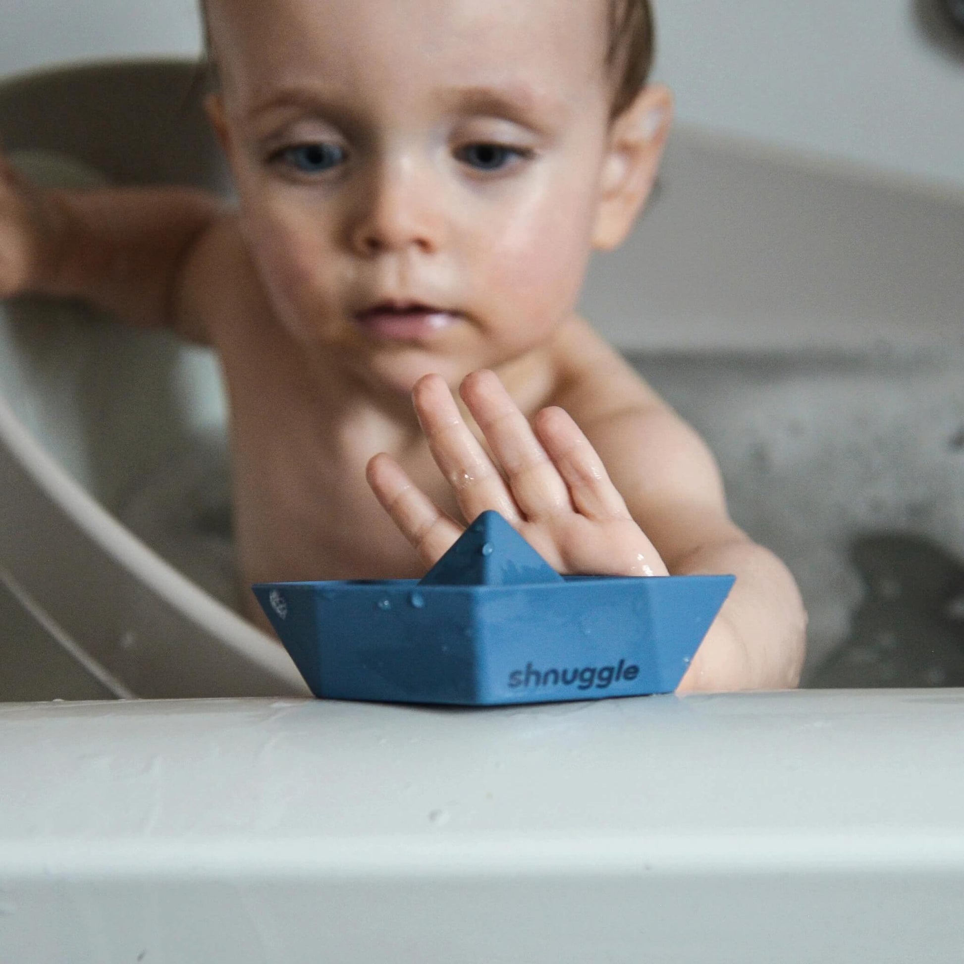 A baby reaching towards a blue bath boat resting on a bath edge, showing scale during use.