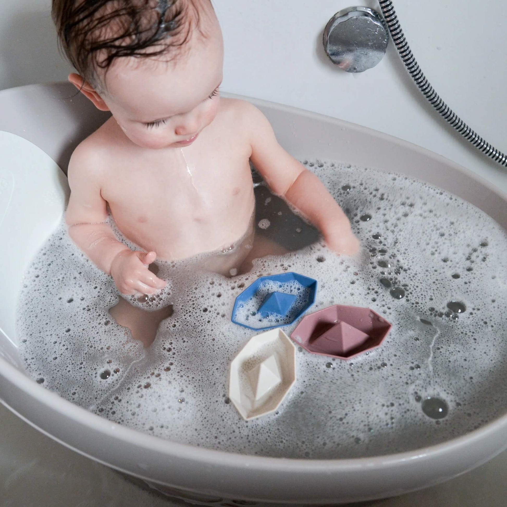 A baby sitting in a bath filled with bubbles, looking at small floating boat toys during bath time.