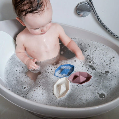 A baby sitting in a bath filled with bubbles, looking at small floating boat toys during bath time.