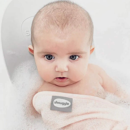 A baby sitting upright in a bath with water and bubbles, wrapped in a white wash cloth with a small stitched fabric label visible on the edge.