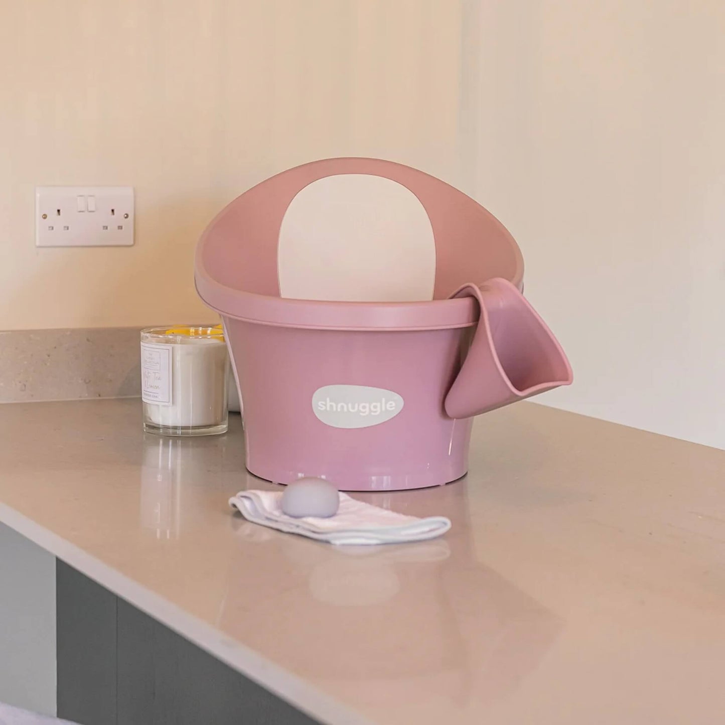 A pink rinsing jug hooked onto the rim of a baby bath on a countertop, showing how it stores neatly within reach between uses.