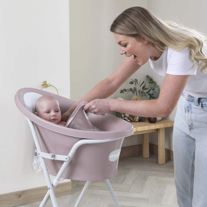 A mother pouring water from a pink rinsing jug over a baby sitting in a raised baby bath, with water directed away from the baby’s face.