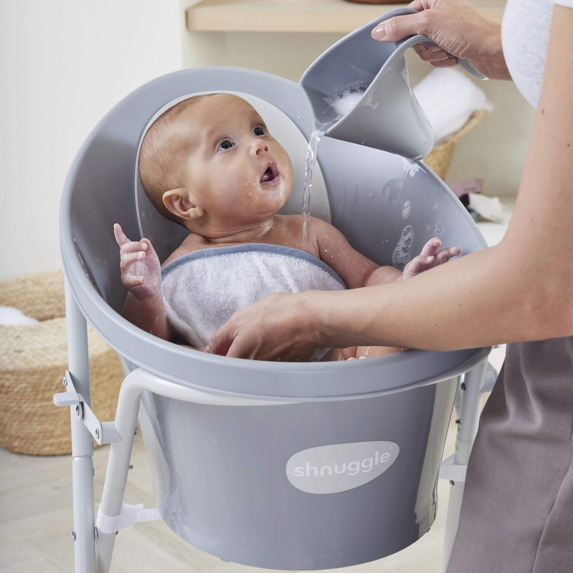 A parent pouring water from a grey rinsing jug over a baby sitting in a raised baby bath, with water directed away from eyes and face.