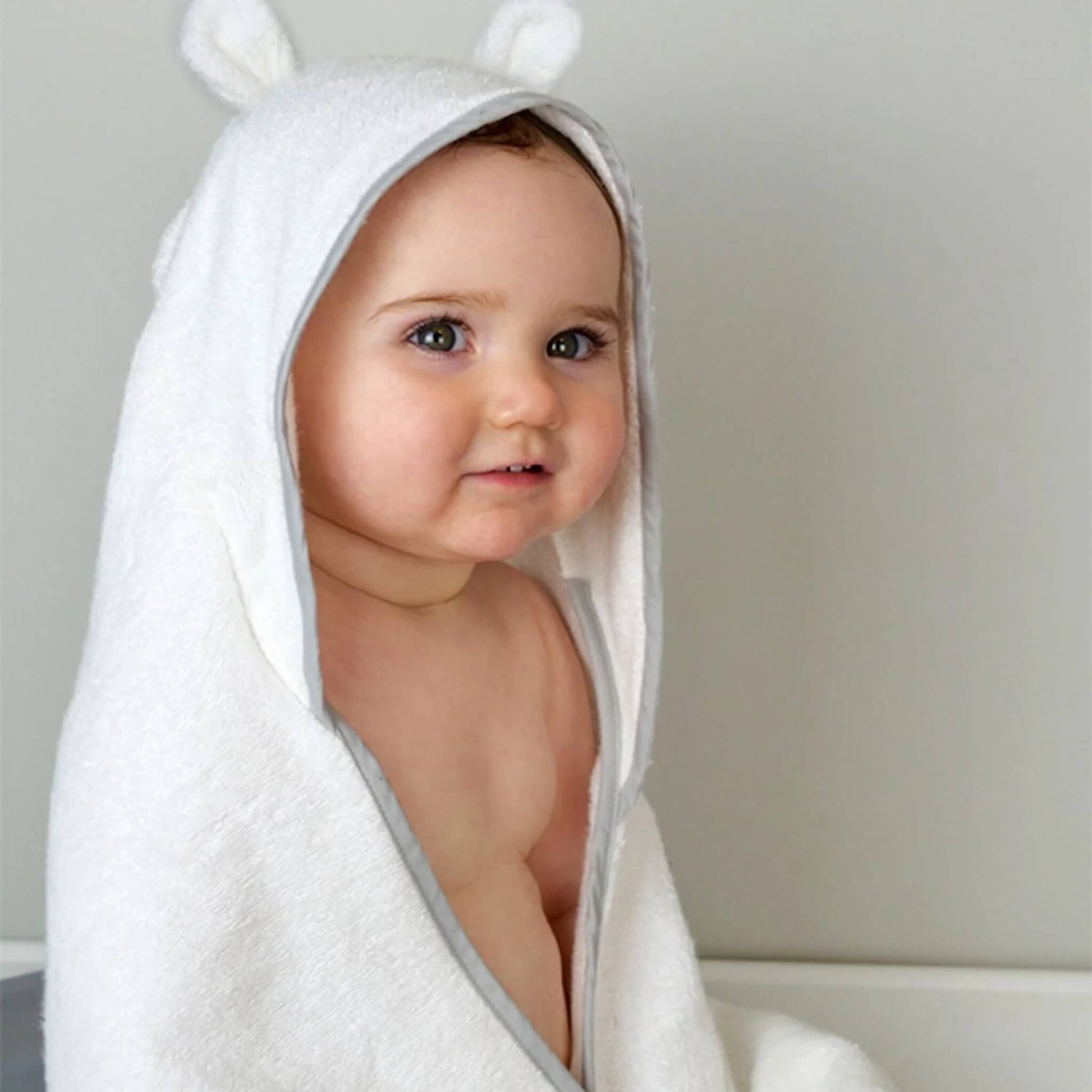 A baby sitting wrapped in a hooded towel with bear ear details, showing a close view of the towel texture and hood shape.