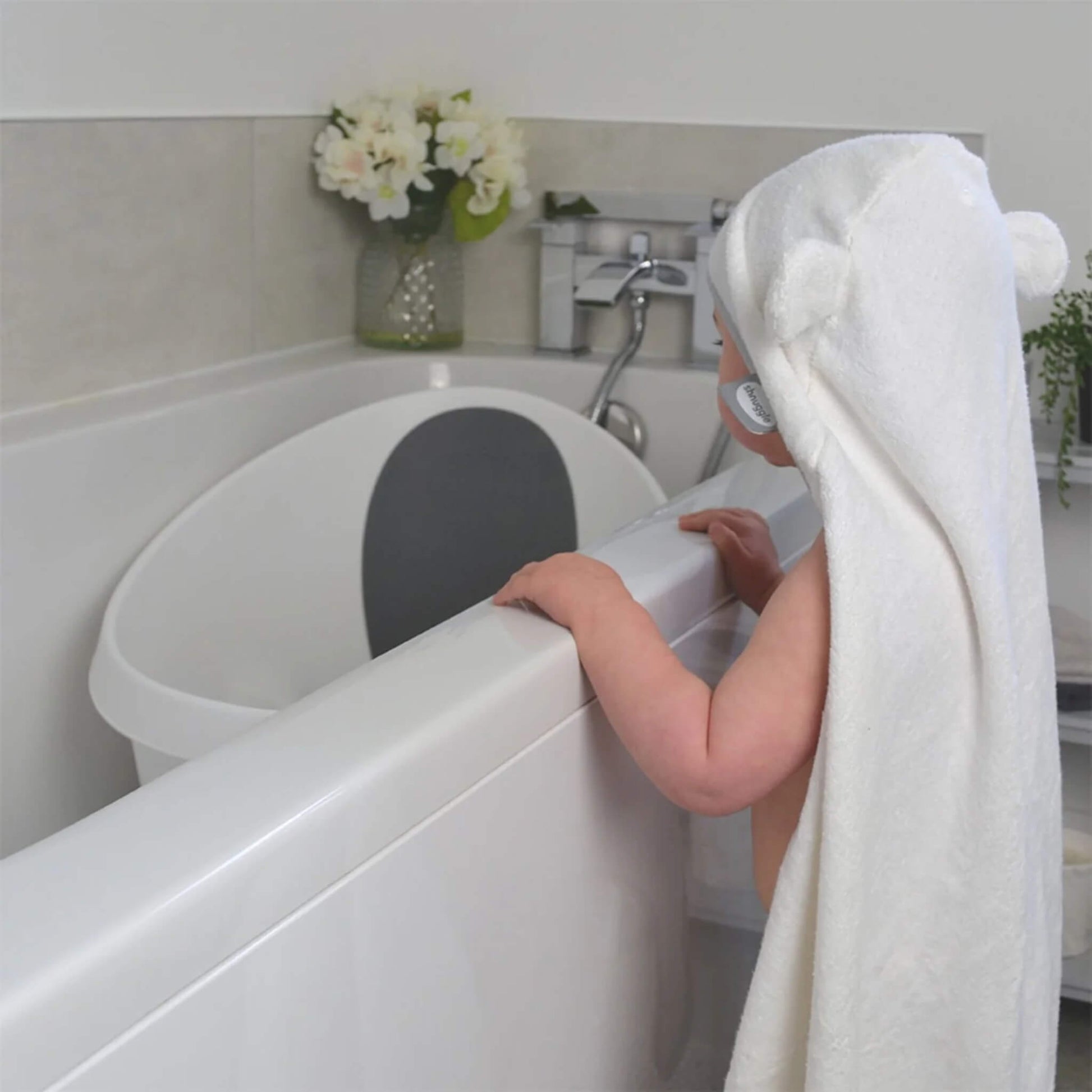 A baby standing at a bath edge while wrapped in a hooded towel, showing the towel length and coverage from behind.