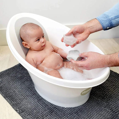 A baby sitting in a white baby bath while an adult holds two star-shaped bath toys above the water.