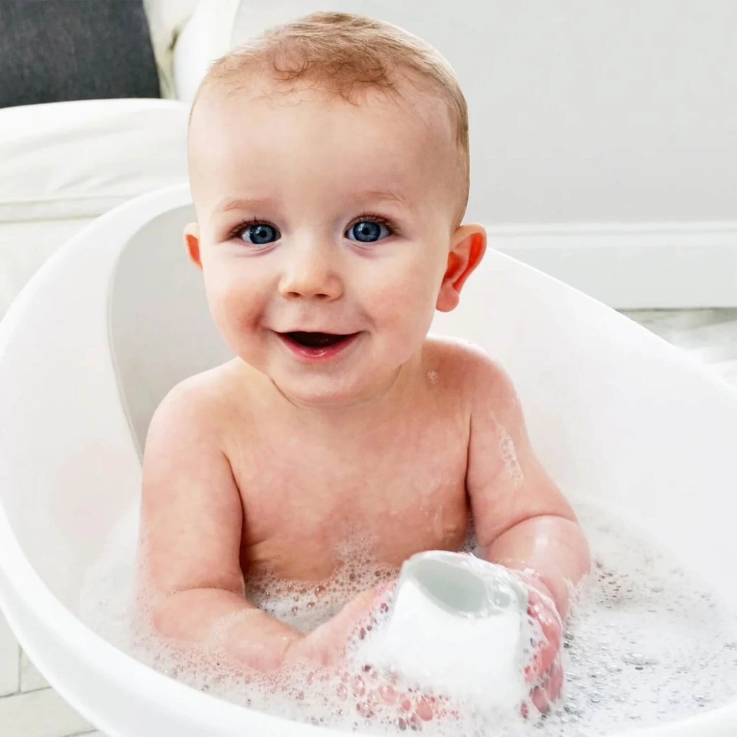 A baby smiling in a white baby bath while holding a star-shaped bath toy filled with water.
