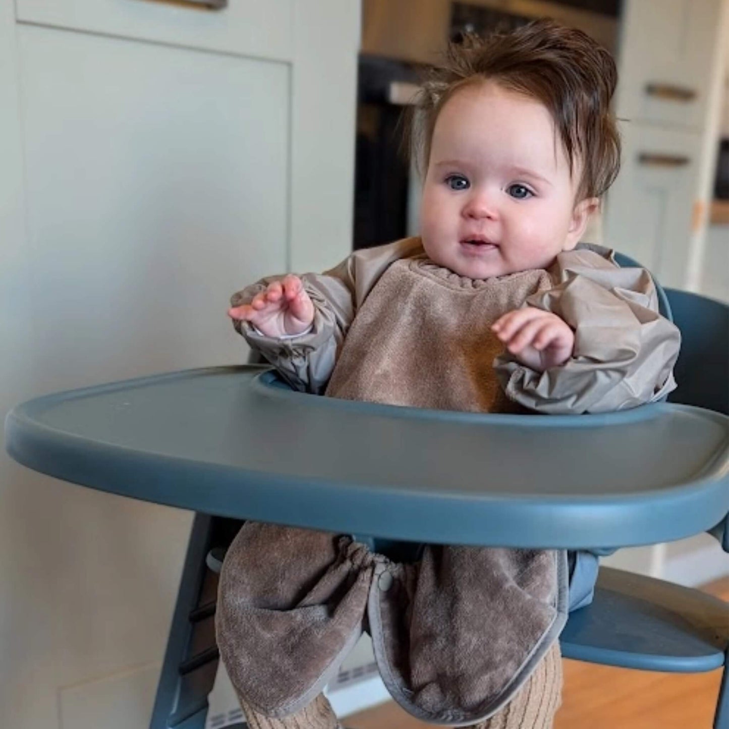 A baby seated in a highchair wearing a taupe long sleeve towel bib, showing the elasticated cuffs and wide coverage across the tray area while sitting upright.