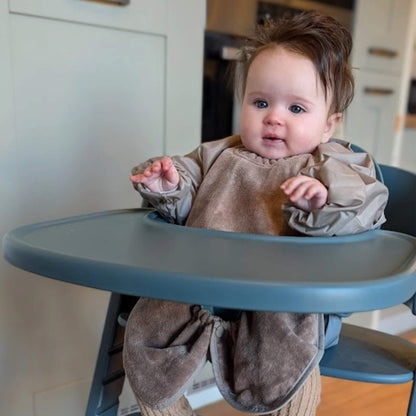 A baby seated in a highchair wearing a taupe long sleeve towel bib, showing the elasticated cuffs and wide coverage across the tray area while sitting upright.