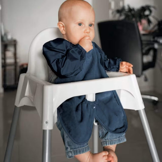 A baby wearing a long sleeve navy highchair bib while sitting in a highchair, showing full arm coverage, soft towel front and elasticated cuffs during feeding.