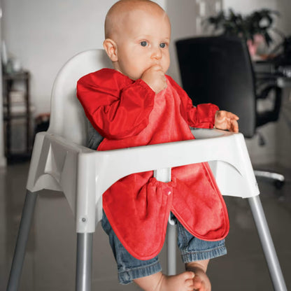 A baby wearing a long sleeve red highchair bib while sitting in a highchair, showing full arm coverage, soft towel front and elasticated cuffs during feeding.