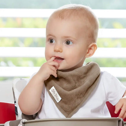 A baby sitting in a highchair wearing a soft taupe bandana-style bib, with one hand raised to the mouth and a blurred window background behind.