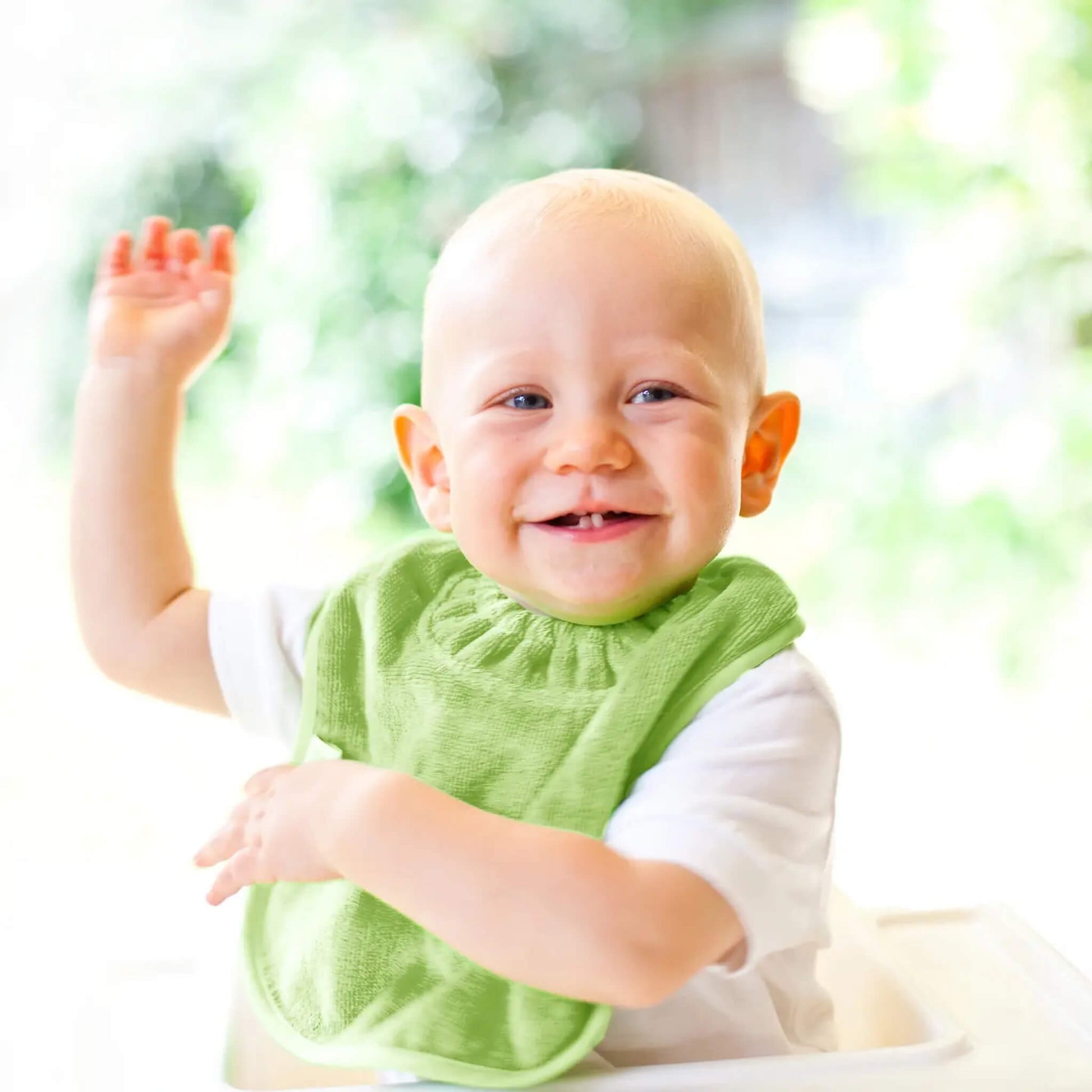 A smiling baby in a highchair wearing a bright sage green towelling bib, raising one arm with blurred greenery in the background.