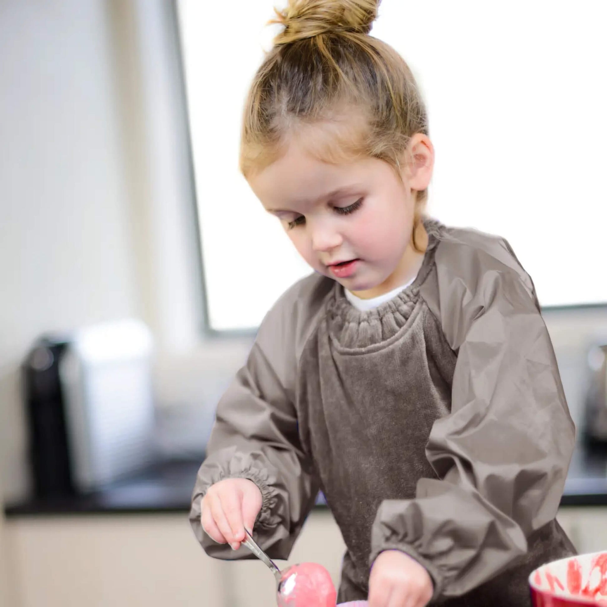 A young child standing at a counter wearing a taupe long-sleeve bib while scooping food from a bowl, with soft natural light in the background.