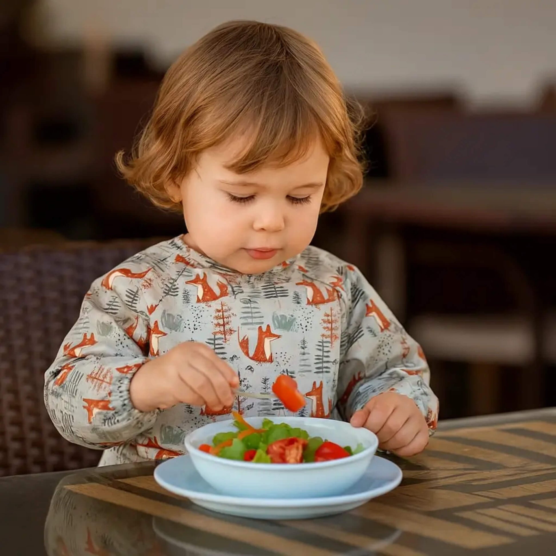 A toddler sitting at a table wearing a fox-pattern long-sleeve bib while picking up salad pieces from a bowl, with warm indoor lighting in the background.