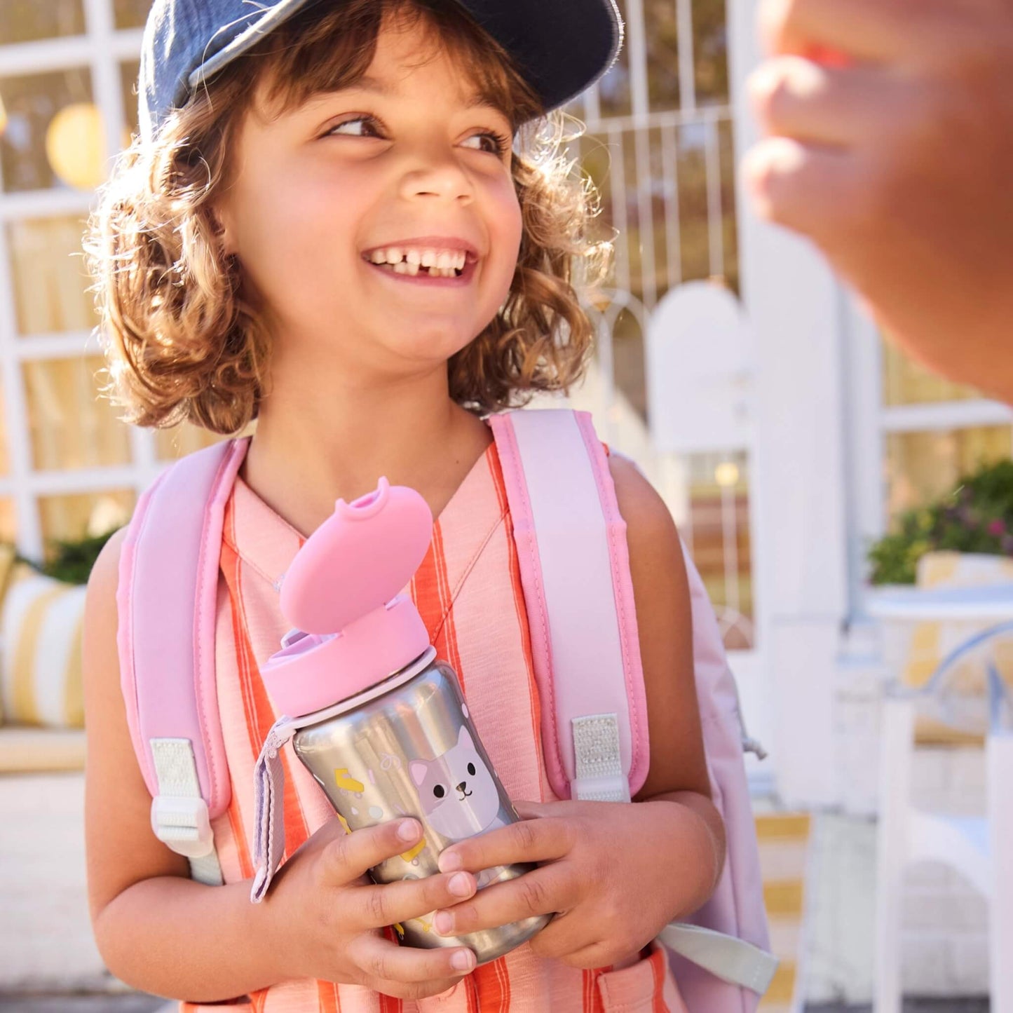 A smiling young girl with curly hair, wearing a blue cap and a pink backpack, holds a stainless steel water bottle with a pink lid in an outdoor patio setting.