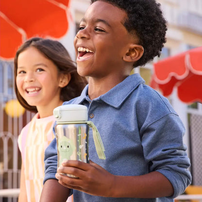 A smiling boy in a blue top holding the dinosaur character stainless steel water bottle, with a smiling girl standing beside him outdoors.