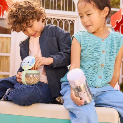 A girl in a turquoise top holds the silver unicorn water bottle while sitting beside a boy using a green elephant snack cup outdoors.