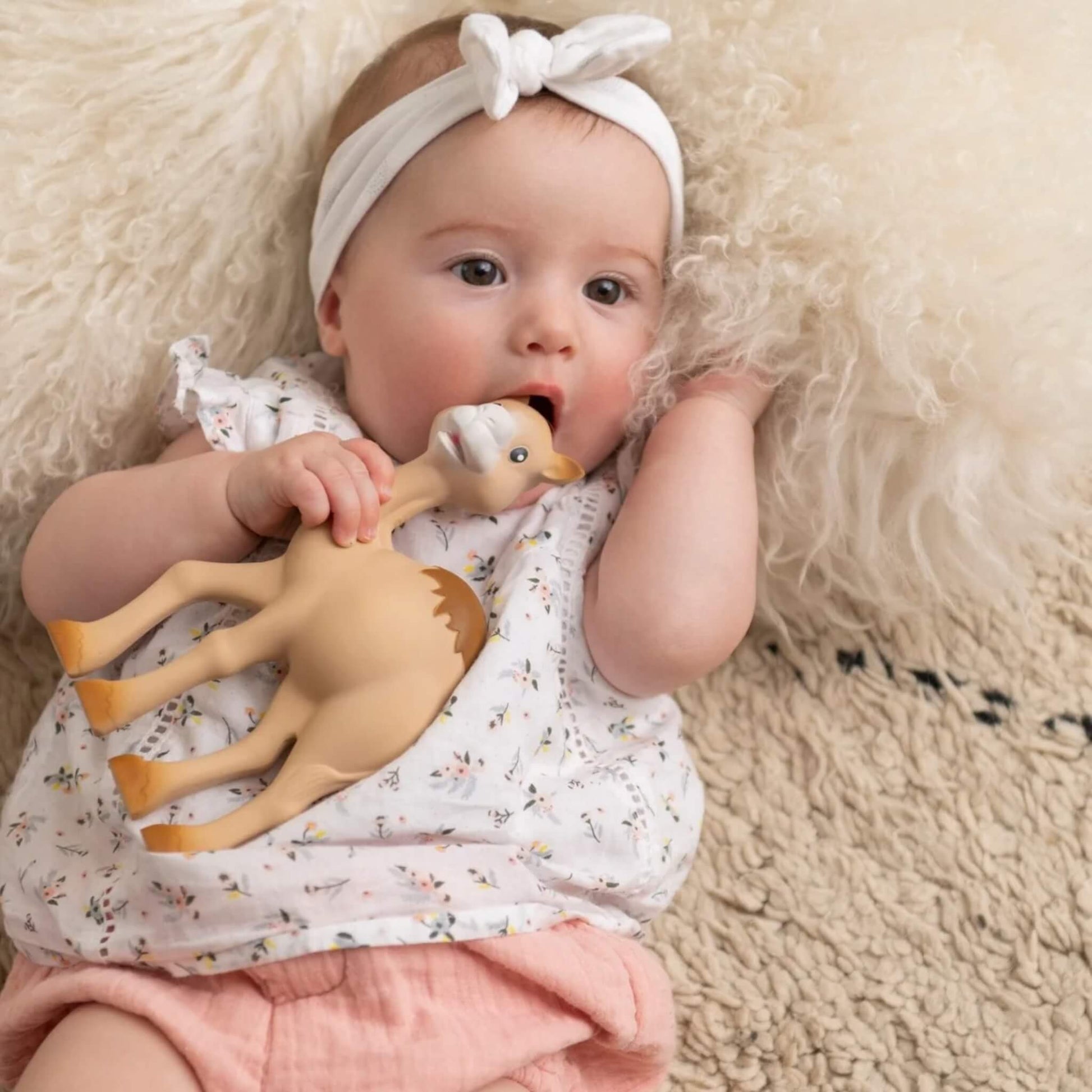 A baby lying on a textured rug, holding and chewing a camel-shaped natural rubber teething toy during calm playtime.