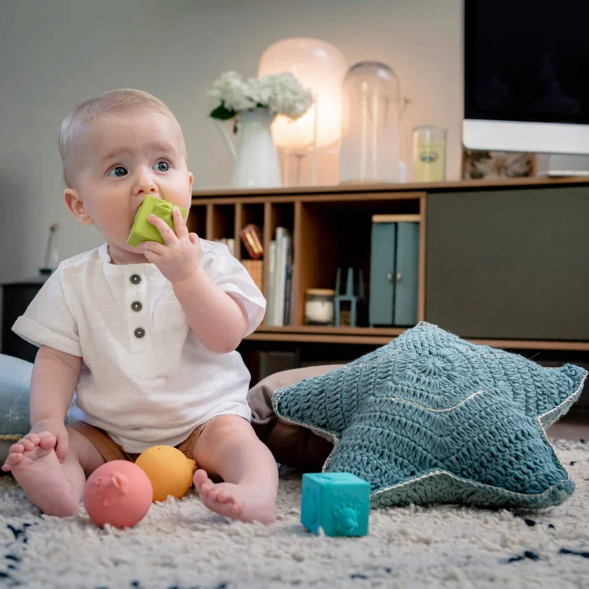 A baby sitting on a rug chewing a green rubber cube, with other textured rubber balls and cubes placed around them during floor play.