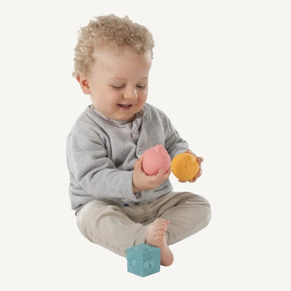 A toddler sitting on the floor holding two soft rubber sensory balls, with textured rubber cubes positioned nearby.