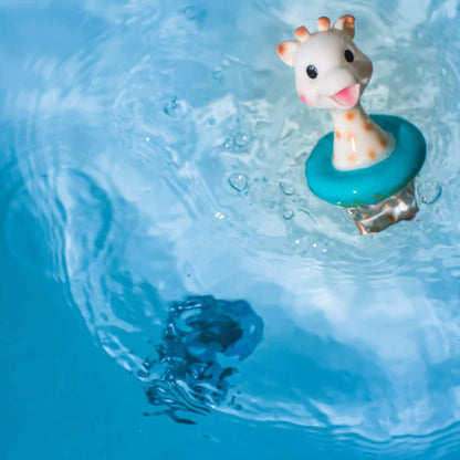 A giraffe bath toy floating upright in blue bath water with ripples around the floating ring.