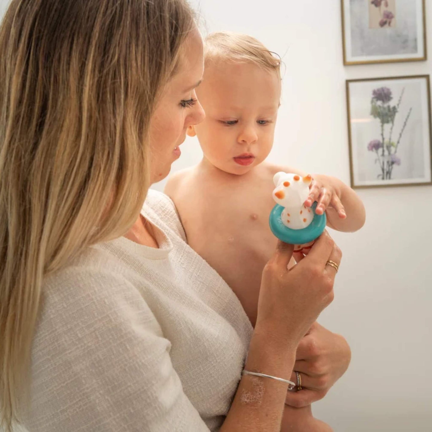 A mother holding a baby during bath time while the baby holds a giraffe-shaped bath toy with a floating ring.