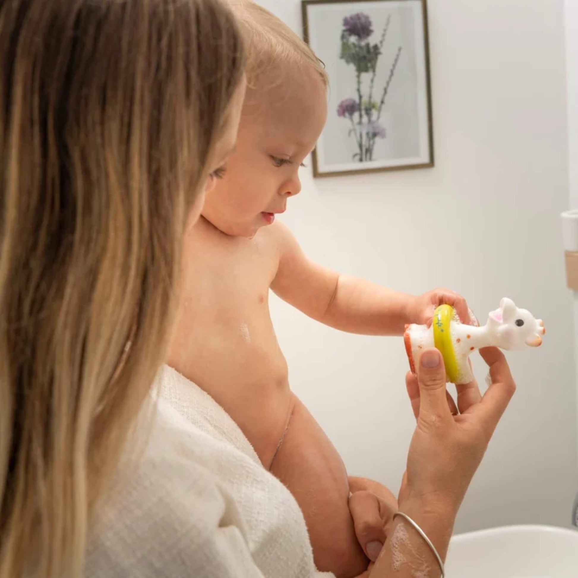 A mother supporting a baby by the bath while the baby reaches towards a giraffe-shaped bath toy held in the hand.
