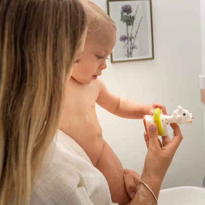 A mother supporting a baby by the bath while the baby reaches towards a giraffe-shaped bath toy held in the hand.
