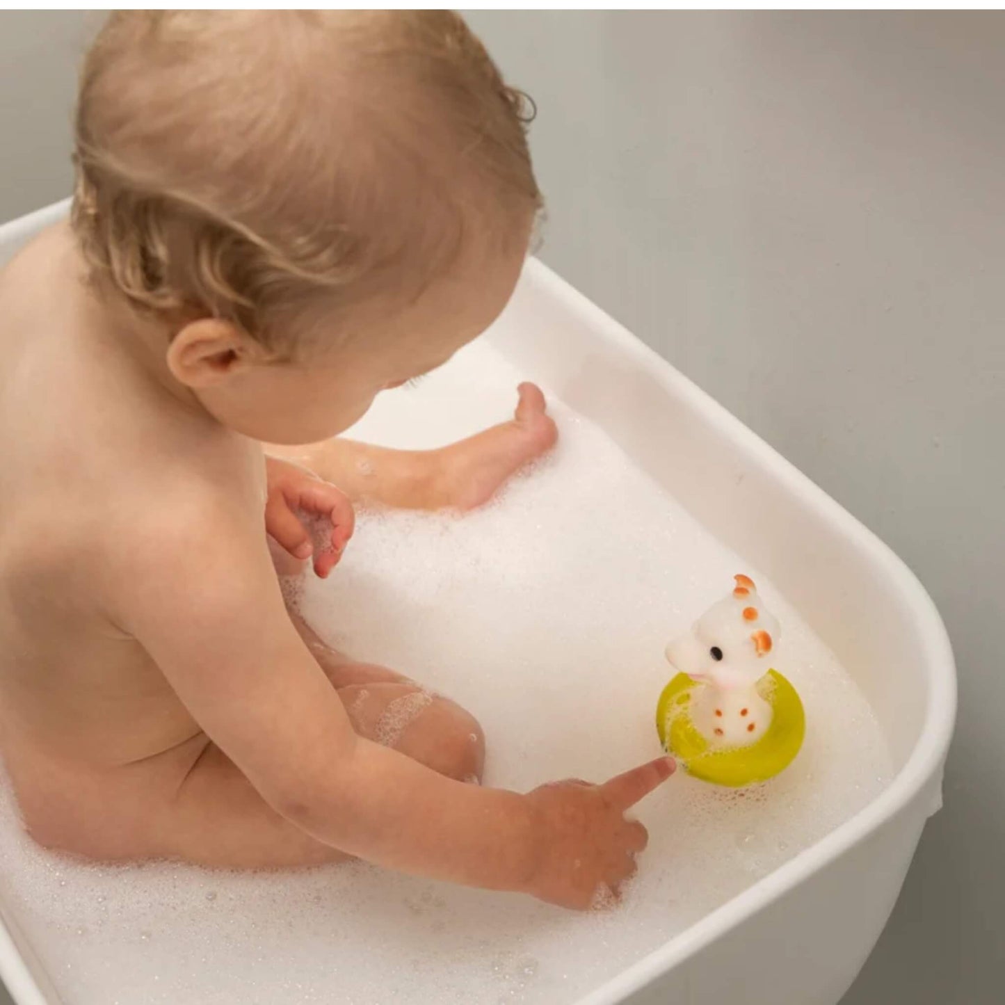 A baby sitting in a white bath with foam, pointing towards a giraffe-shaped bath toy floating nearby.