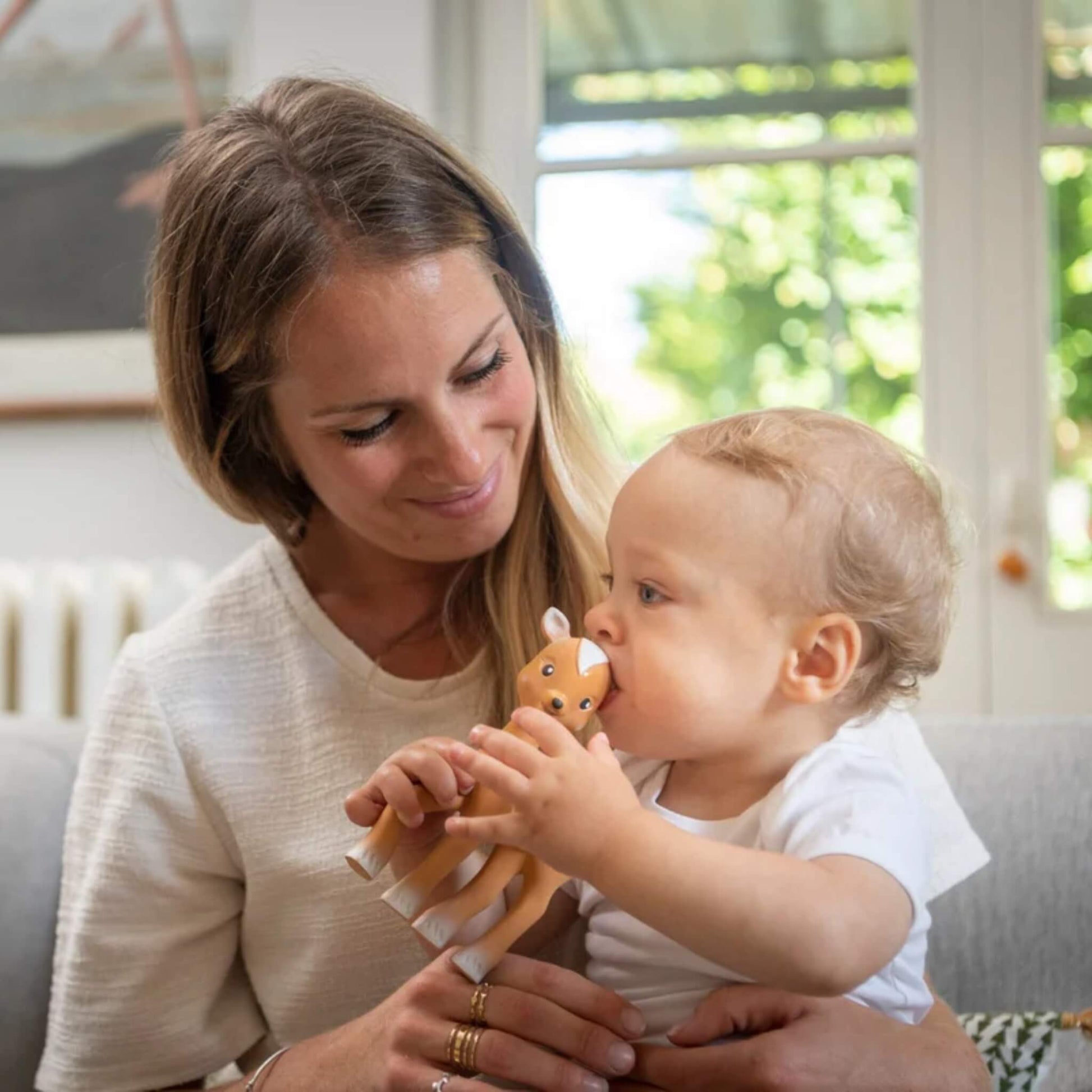 A mother holding a baby who is chewing a fawn-shaped natural rubber teething toy while sitting together in a bright living room.