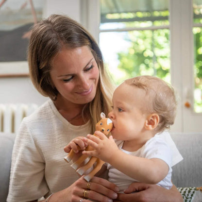 A mother holding a baby who is chewing a fawn-shaped natural rubber teething toy while sitting together in a bright living room.
