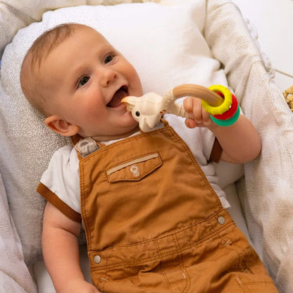 A baby lying on cushioned bedding while chewing a giraffe-shaped natural rubber rattle teether with colourful textured rings.