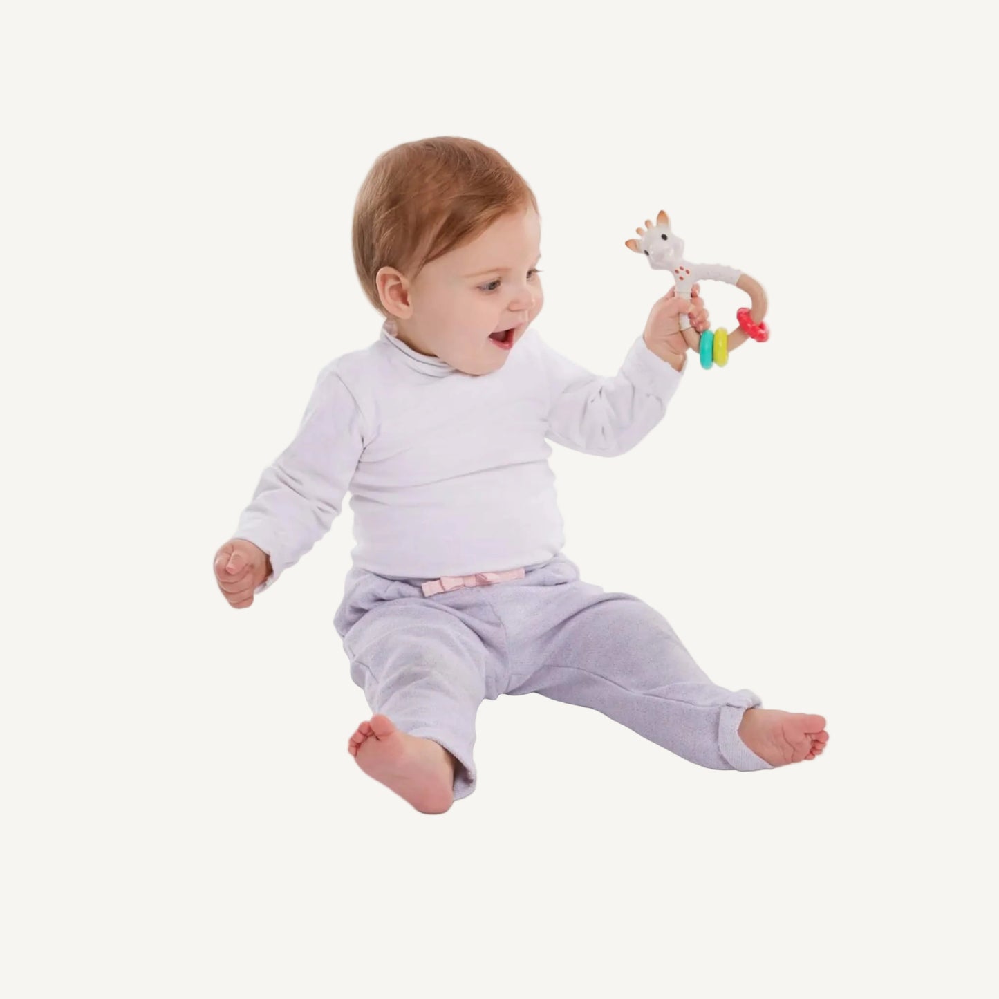 A baby sitting upright while holding and examining a giraffe-shaped natural rubber rattle teether with three textured rings.