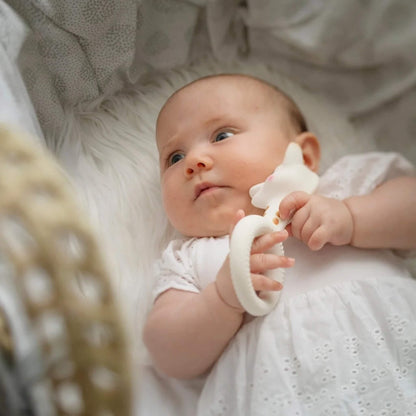 A baby lying on soft bedding while holding a giraffe-shaped natural rubber ring teether close to the face.