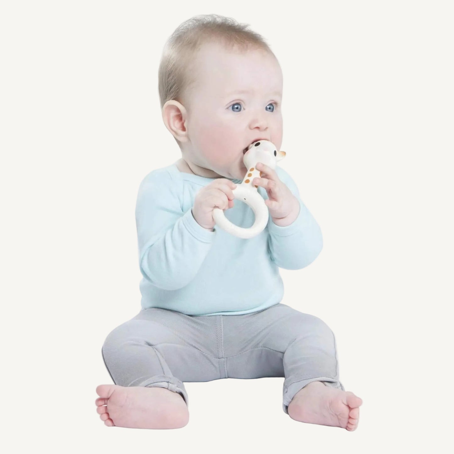 A baby sitting upright while chewing a giraffe-shaped natural rubber ring teether held with both hands.