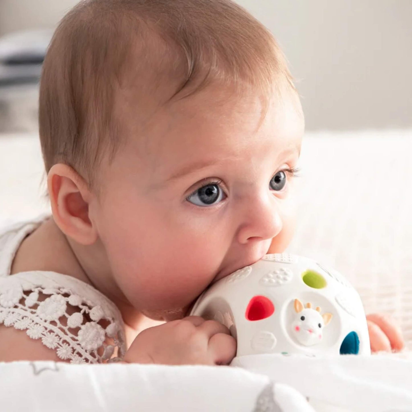 Baby lying on soft bedding chewing a textured sensory ball with colourful sections and raised patterns for teething relief.