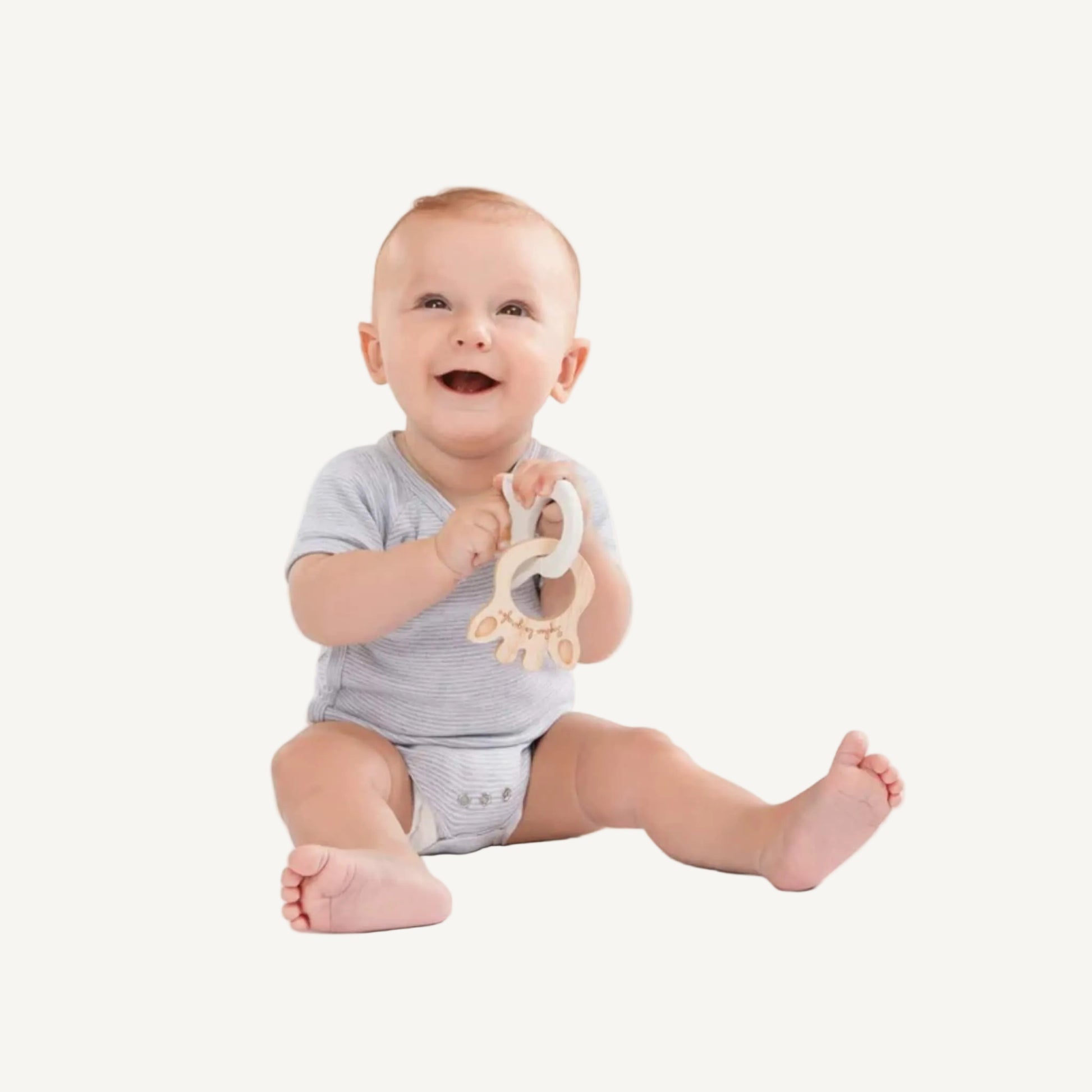 A smiling baby sitting upright while holding a giraffe silhouette teething ring made from rubberwood and natural rubber.