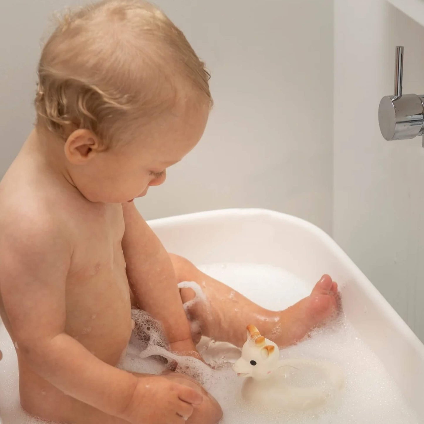 Baby sitting in a white bath filled with bubbles, reaching toward a floating giraffe bath toy.