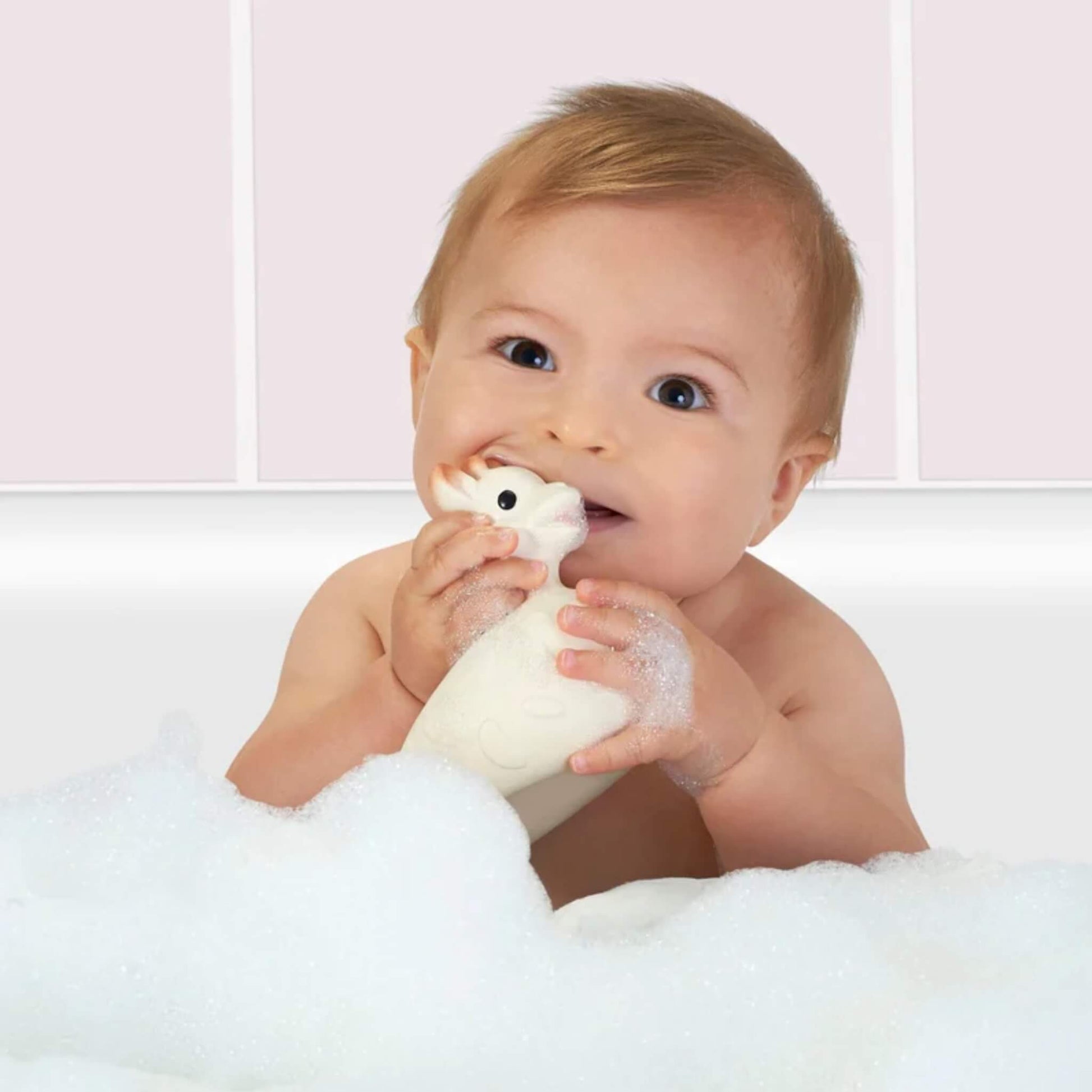 Baby sitting in a bath and holding a rubber bath toy surrounded by bubbles.