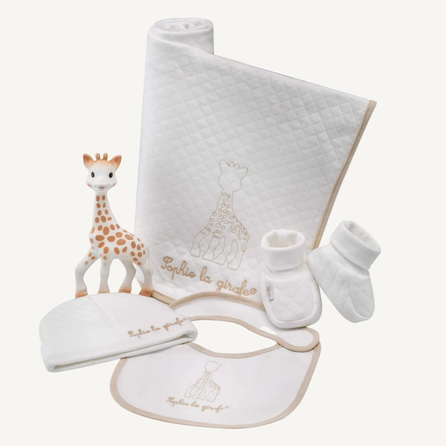 A coordinated newborn gift set laid out flat, showing a white quilted blanket, matching hat, bib, booties, and a natural rubber giraffe toy.