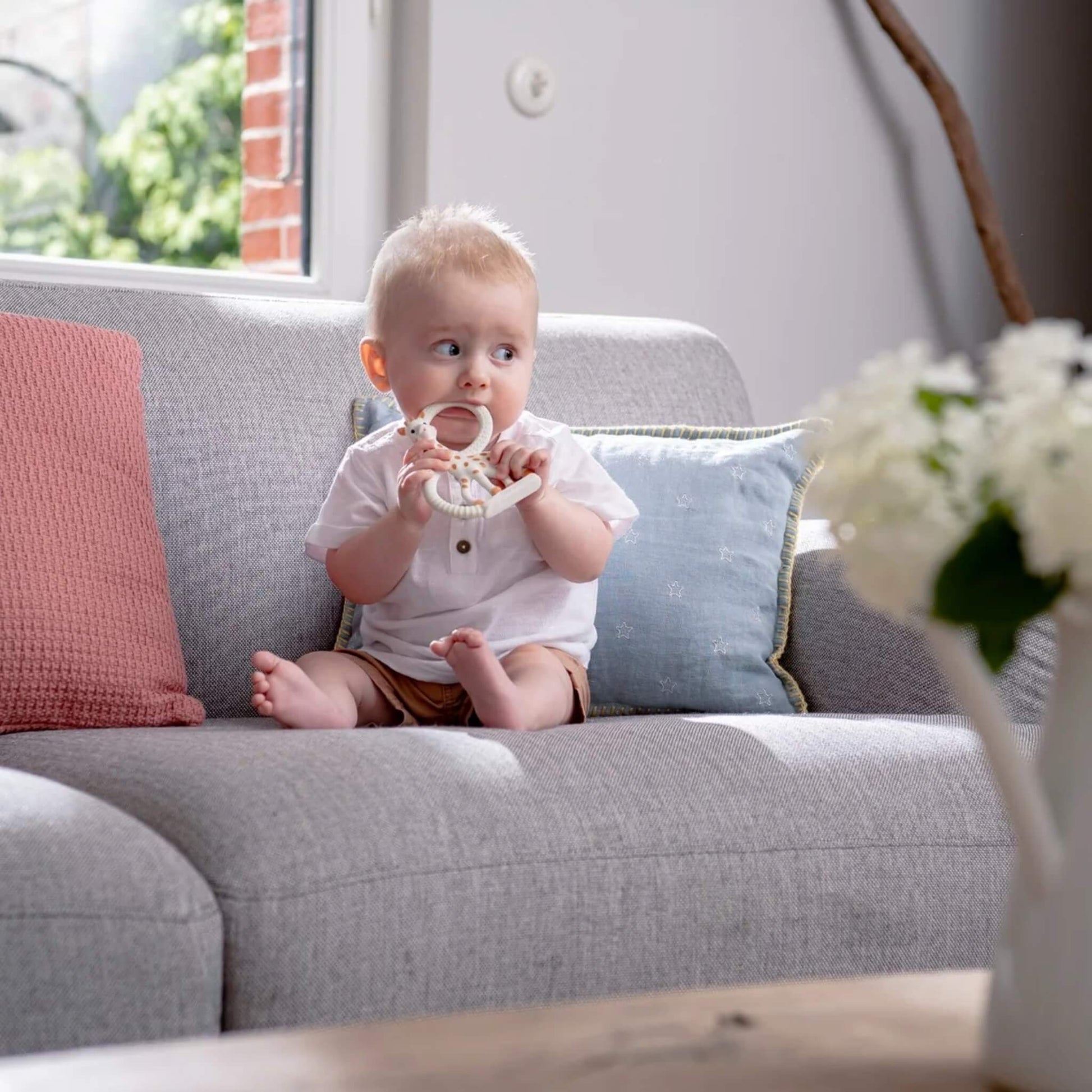 A baby sitting on a sofa while chewing a giraffe-shaped natural rubber teething ring during calm indoor play.