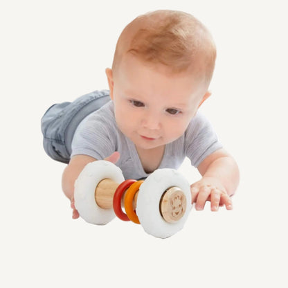 A baby lying on a play surface while reaching forward to roll a wooden and natural rubber rattle toy.