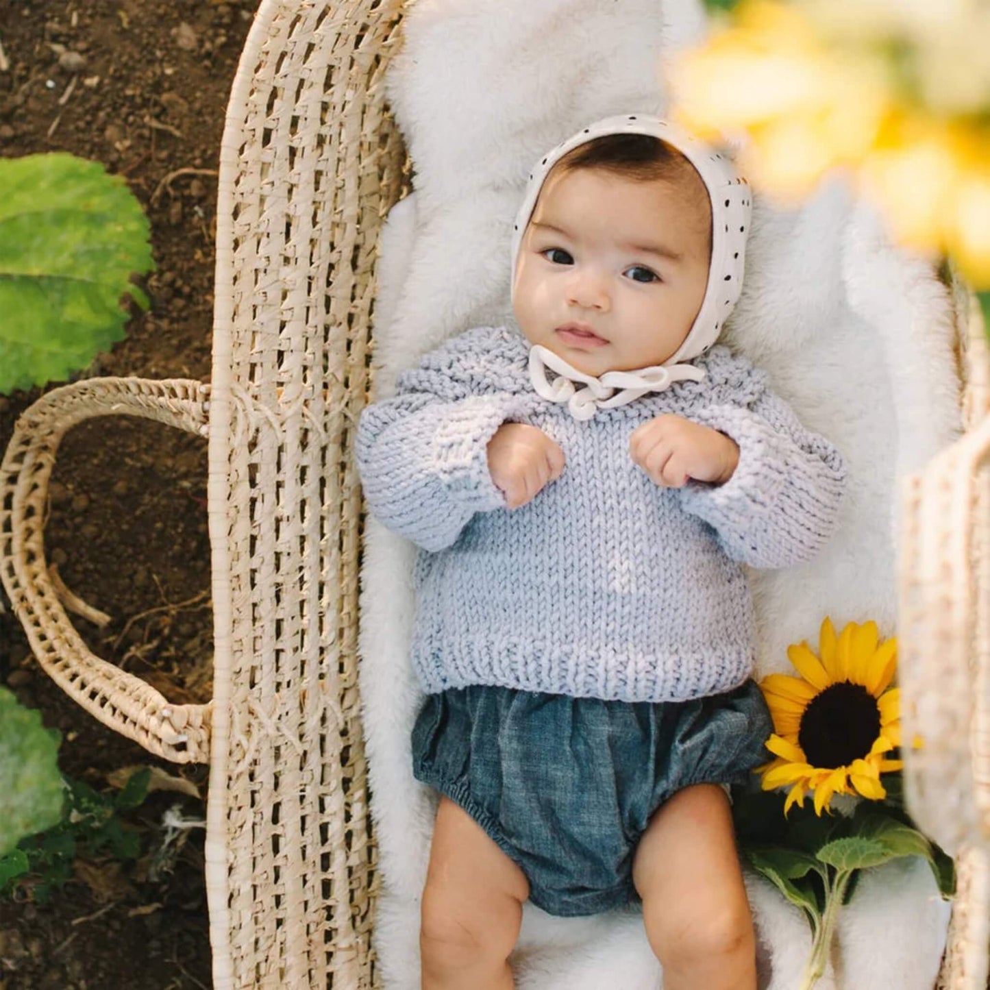 A baby lying in a woven basket wearing a hand-knitted pale blue sweater with ribbed cuffs, paired with soft bloomer shorts and a bonnet, photographed outdoors among leaves and flowers.